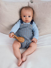 Load image into Gallery viewer, Baby in checkered overalls holding a wooden toy on a beige couch
