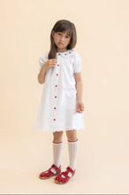 Load image into Gallery viewer, Young girl wearing a white dress with red buttons on a beige background