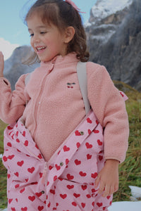 Child wearing a pink fleece jacket and red heart-patterned pants outdoors with mountains in the background