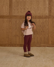 Load image into Gallery viewer, Child wearing a pink shirt, maroon pants, and a matching hat against a wooden paneled wall.