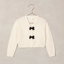 將圖片載入圖庫檢視器 An off-white knit cardigan sweater with two black bow details on the front, hanging on a metal hanger against a textured beige background.