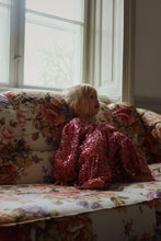 Load image into Gallery viewer, Child in a red sequin dress sitting on a floral couch by a window