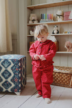 Load image into Gallery viewer, Child in red pajama set standing in a room with shelves and toys.