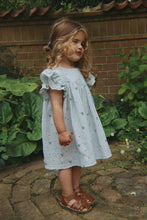 Load image into Gallery viewer, Young girl in a light blue dress standing outdoors near a brick wall and greenery.