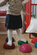 Load image into Gallery viewer, A child wearing a leopard print top, dark plaid skirt, and white knit tights balances on a red, green, and blue textured foam dome on a wooden floor in a nursery.